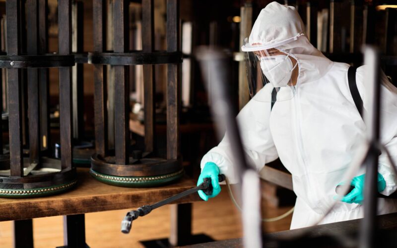 Disinfection worker in hazmat suit spraying contaminated cafe due to coronavirus epidemic. Sanitation worker in protective suit disinfecting empty cafe during COVID-19 epidemic.