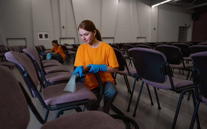 Two cleaning company workers sanitizing the chairs in sitting area Attentive young woman vacuum-cleaning upholstery on chairs with the help of her collegue